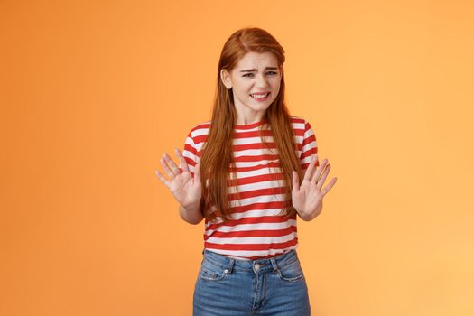 Reluctant bothered cute redhead girl apologizing make stop refusal sign, hold hands block, grimacing, cringe unwilling participate, rejecting offer, stand orange background uncomfortable, step back.