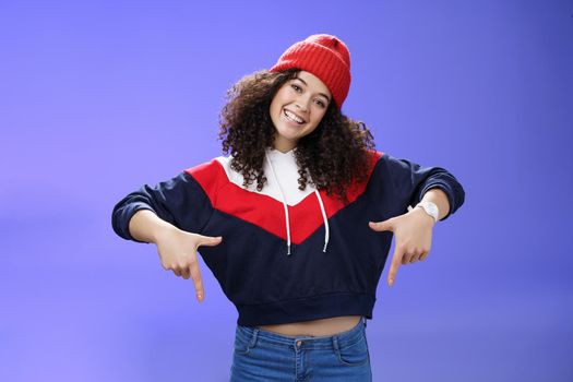 Waist-up shot of cool and stylish young woman with curly hair smiling flirty and joyful pointing down showing promotion as tilting head happily and posing over blue background in outdoor clothes.