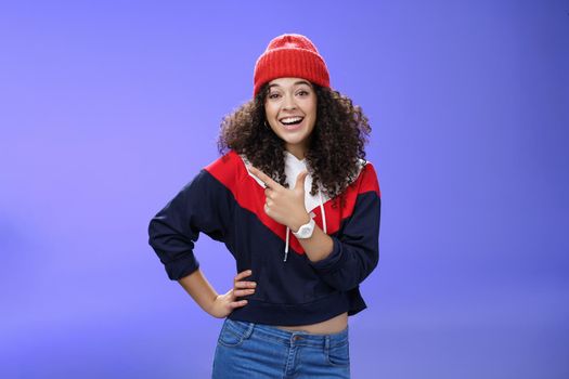 Stylish and sociable friendly-looking attractive woman with curly hair in warm beanie pointing at upper left corner laughing from joy and amazement having fun posing happily over blue background.