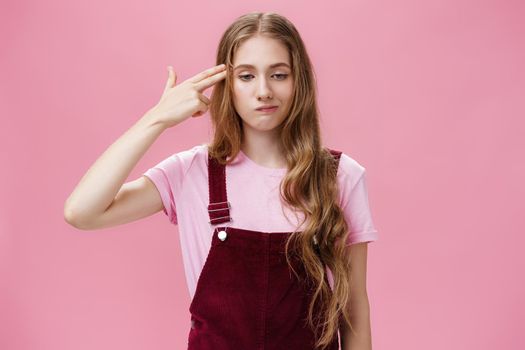 Portrait of bothered uneasy cute european female with long natural fair hair in trendy corduroy overalls smirking from irritation and annoyance showing finger fun gesture as if shooting in head. Copy space