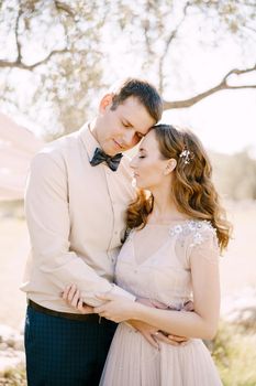 Groom hugs bride while standing under the tree. Portrait. High quality photo