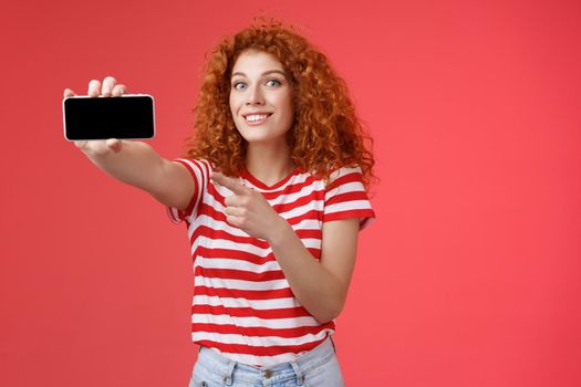 Excited happy good-looking redhead curly female show horizontal smartphone screen pointing display gadget smiling pleased proud beat friends score game standing red background. Technology concept
