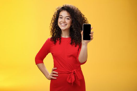 Girl brag with new phone she got on christmas feeling delighted holding mobile device in hand showing smartphone screena t camera, smiling broadly with uplifted mood over yellow background. Technology concept