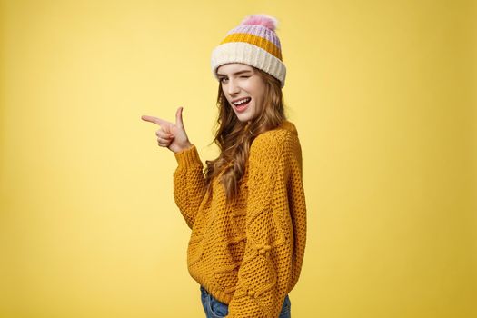 Profile shot sassy flirting young cute woman winking seduce you smiling cheeky turn camera pointing left index finger showing copy space suggest go drink coffee together, standing yellow background.