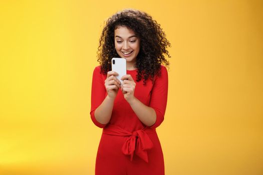 Wow new mobile phone amazing. Impressed and astonished good-looking curly-haired female in red dress holding smartphone looking at screen amused as playing cool app or game over yellow wall.