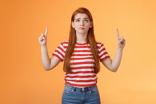 Hesitant upset cute redhead woman weighing choices look doubtful uncertain, frowning thoughtful, ponder decision, look pointing up unsure, have doubts, stand insecure orange background.