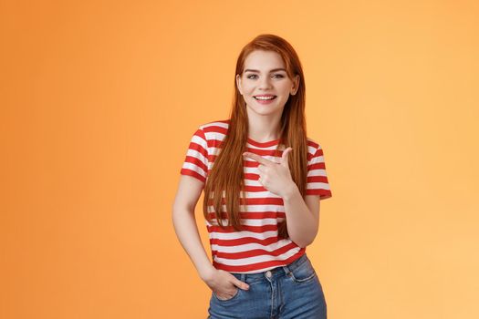Carefree modern 20s female student chatting friends discuss left copy space, pointing left relaxed casual pose, smiling camera joyfully, introduce advertisement product, orange background.