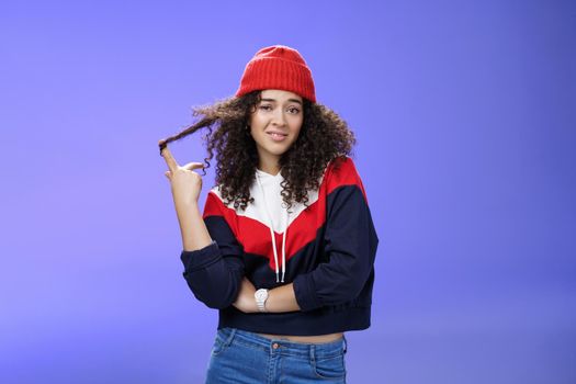 Stylish and snobbish arrogant curly-haired woman in warm beanie rolling curl on finger smirking and looking with contempt at camera, scorning person as being too cool over blue background.