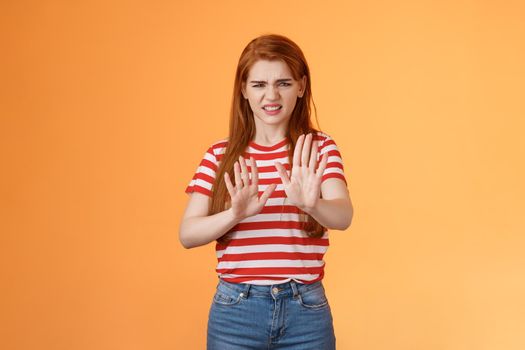 Gosh it stinks. Disgusted redhead picky woman blocking sign raise hands up defensive, grimacing, cringe from aversion awful smell, show refusal rejecting disgusting offer, stand orange background.