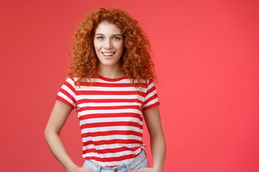 Sassy confident cheeky good-looking redhead curly-haired woman hold hands pockets smiling daring assertive motivated have perfect summer days wear striped trendy t-shirt stand red background.