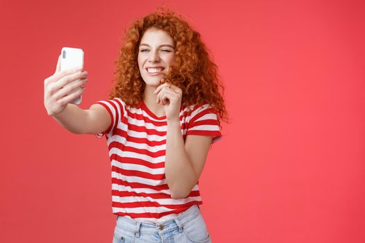 Sassy good-looking stylish charismatic redhead female curly hairstyle winking cheeky expression making flirty kinky faces hold smartphone taking selfie record video message play funny facial filters.