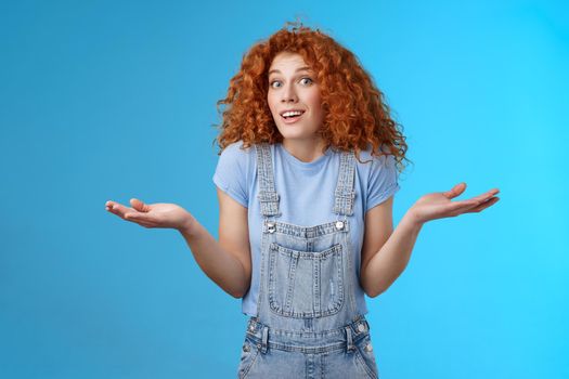 Who knows. Careless clueless attractive playful redhead cute curly-haired girl shrugging unaware hold hands sideways unaware smiling act innocent uninvolved not knwo answer blue background.