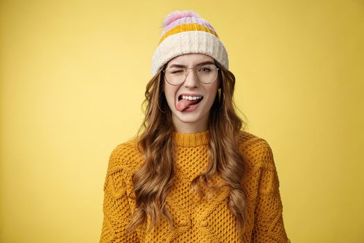 Carefree attractive funny happy caucasian girl 20s curly-haired wear glasses hat sweater having fun playfully show tongue, winking flirty expressing positive optimistic mood, yellow background.