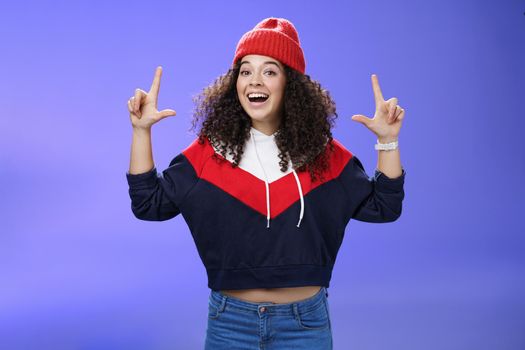 Waist-up shot of cheerful energized young woman calling friend inviting hang out as weather awesome pointing up with raised hands smiling broadly posing in red winter hat over blue background.