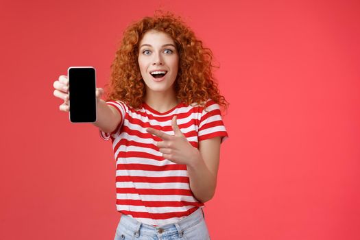 Take look what awesome smartphone. Impressed excited good-looking female love digital innovations redhead girl curly hairstyle showing phone screen pointing display promote cool app.