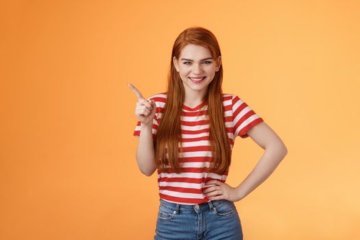 Cheerful sly redhead girlfriend have something interesting on mind, squinting cunning joyful smile, hold hand waist confident pose, pointing upper left corner give recommendation, orange background.