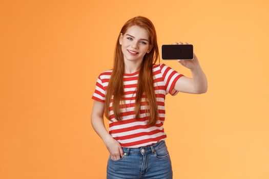 Sassy confident attractive 20s caucasian ginger girl introduce smartphone app, recommend cool game, show cellphone screen horizontal, smiling proud, brag beating score, stand orange background.