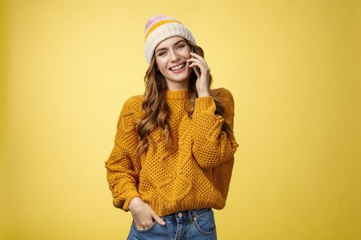Charming stylish european woman wearing hat sweater tilting head flirty smiling talking smartphone calling boyfriend consulting friend via cellphone, standing happily casual pose yellow background.