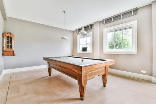 A room with a large wooden pool table and several windows on the wall