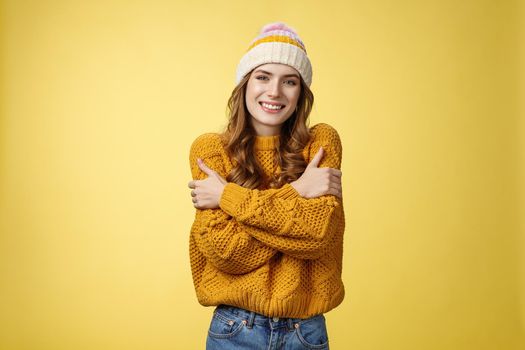 Getting cold bring jacket. Portrait charming tender feminine cute girlfriend embacing herself cuddling smiling warm gaze freezing outside standing knitted sweater hat, yellow background.