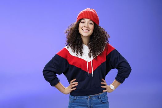 Confident ad energized cute european woman with curly hairstyle in warm beanie and stylish sweatshirt holding hands on waist and smiling friendly at camera as ready to go out and have fun outside.