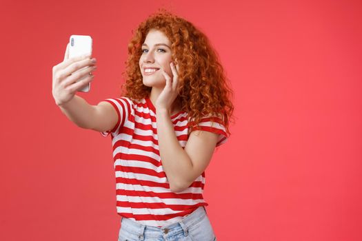 Popular cheerful good-looking stylish female blogger redhead curly hairstyle feel pretty self-acceptance taking selfie raised arm holding smartphone posing silly cute phone camera red background.