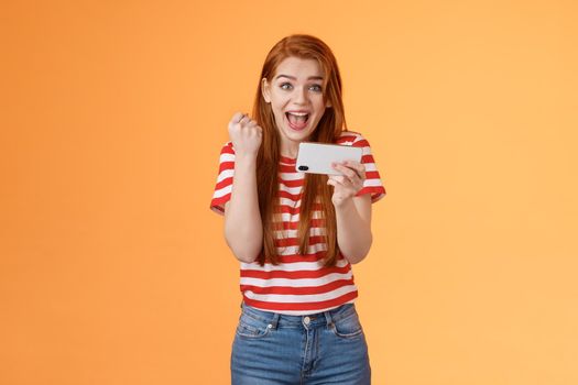 Happy excited redhead female pass level, like awesome game, score goal, hold smartphone horizontally, fist pump success, celebrate lucky achievement, smiling broadly, orange background.
