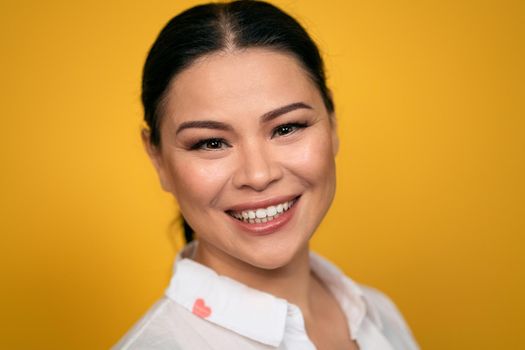 Pleasant looking glad Asian woman looks in camera, has toothy smile, wears casual clothing, isolated on yellow background. Human emotions, facial expression concept. Facial expressions, feelings.