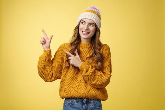 Curious attractive young 20s woman intrigued what hiding left pointing sideways looking interested smiling excited peeking cool fantastic promotion, standing joyfully yellow background. Copy space