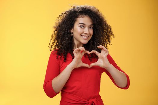 Waist-up shot of romantic and cute pretty girlfriend with curly hair in red dress showing heart sign over chest and smiling broadly confessing in admiration and love posing over yellow background.