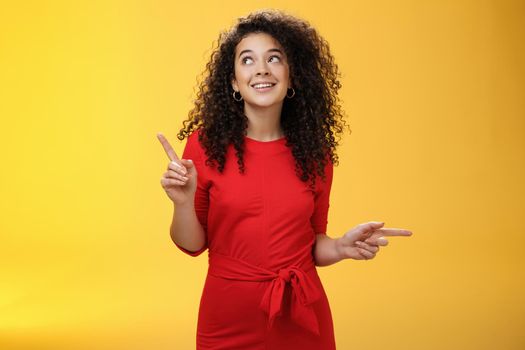 Dreamy charming woman making choice as checking out two awesome products pointing sideways looking intrigued and curious at upper left corner, smiling dreamy and delighted over yellow wall. Body language and advertisement concept