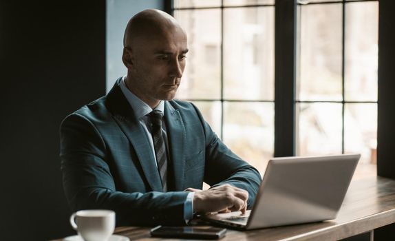 Bald middle aged businessman in business suite working on laptop next to open windows in modern office. Handsome bold man working on laptop in office.