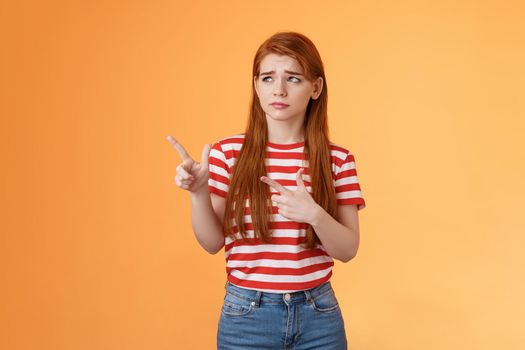 Complicated troubled cute young redhead girl puzzled facing tough difficult decision, look uneasy frowning perplexed hesitating, stare pointing upper left corner disappointed, orange background.