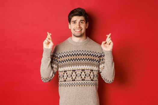 Image of hopeful and worried man in christmas sweater waiting for something, cross fingers for good luck and making wish, nervous about new year gift, red background.