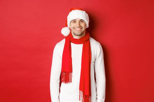 Happy caucasian guy celebrating christmas and smiling, wearing santa hat, scarf and white sweater, enjoying new year holidays, standing over red background.