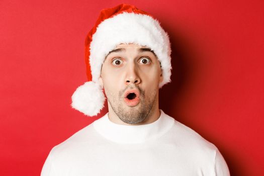 Close-up of attractive surprised man in santa hat, saying wow and looking at something interesting, standing against red background.