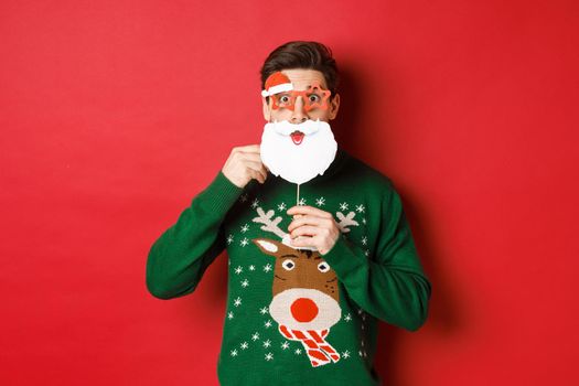 Portrait of surprised man in green christmas sweater, holding funny santa claus mask, celebrating new year, standing over red background.