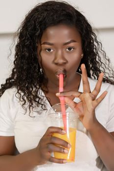 African Model In White T-shirt Drinks an Orange Juice From a Straw. Afro Model Has a Glass of Orange Juice in Her Hand. Close-up. White Background. High quality photo