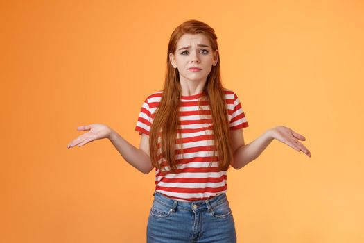 Uneasy questioned and clueless lovely modern stylish redhead woman shrugging spread hands sideways unaware, grimacing perplexed, tough question, have no answer, stand orange background.