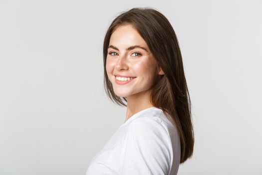 Portrait of confident beautiful brunette woman turning face at camera with dreamy look, smiling over white background.
