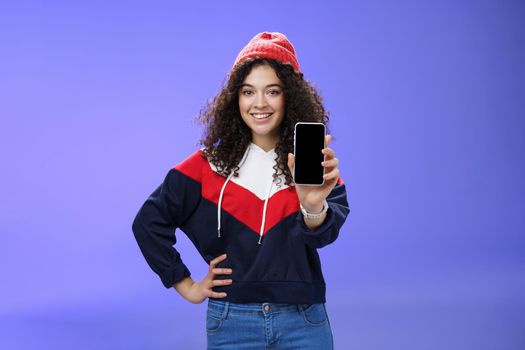 Girl showing her post as pulling smartphone screen towards camera smiling delighted waiting opinion, holding hand on waist in confident pose wearing warm winter red beanie and sweatshirt. Technology and people lifestyle concept