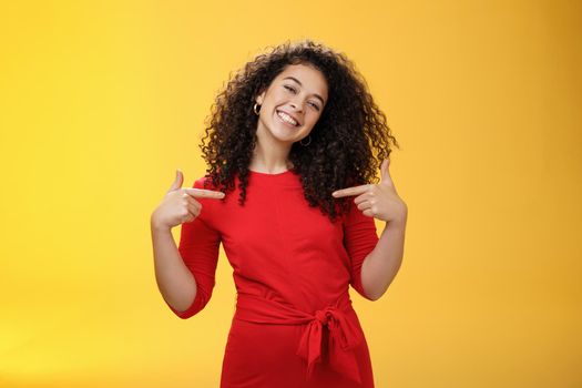Proud and satisfied ambitious successful female student in red dress standing pleased smiling and pointing at herself as if bragging about own achievements happily and glad over yellow wall.