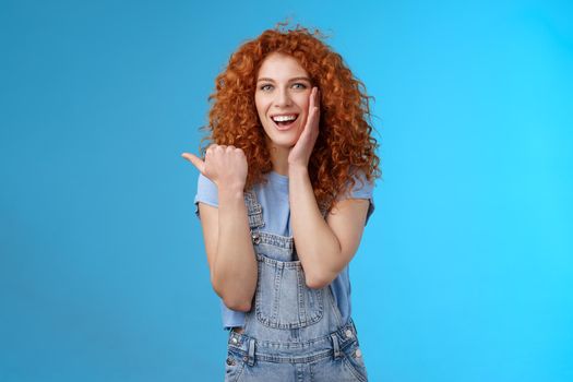 Entertained charismatic good-looking ginger girl curly hairstyle touch cheek gently pleased smiling broadly enthusiastic pointing left thumb impressed excited see awesome promo stunning location.