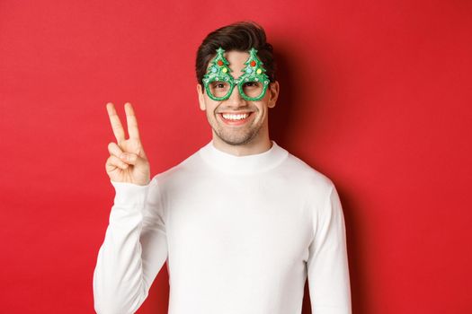 Image of handsome man in white sweater and party glasses, showing peace sign and smiling, wishing merry christmas, standing over red background.
