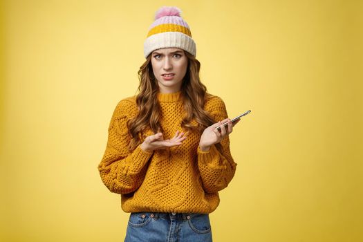 Confused cringing annoyed young woman shocked how friend talked her frowning raise hand dismay look frustrated camera holding smartphone finish video call unpleasant note, yellow background.