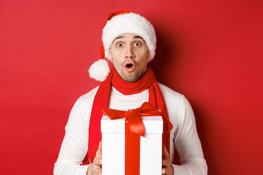 Concept of winter holidays, christmas and lifestyle. Close-up of surprised handsome guy in santa hat and scarf, looking amazed and holding new year gift, standing over red background.