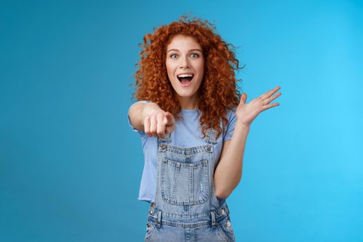 Excited impressed surprised cute redhead european woman curly astonished happily pointing index finger camera gesturing lively thrilled see awesome thing reacting amazed standing blue background.