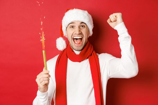 Concept of christmas, winter holidays and celebration. Handsome man celebrating new year and having fun, holding sparkler and smiling, wearing santa hat, standing over red background.