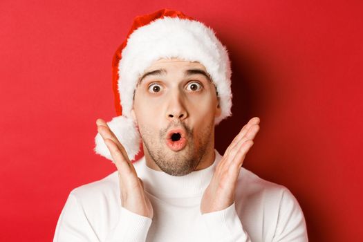 Close-up of amazed handsome man in santa hat and white sweater, raising hands and looking impressed, standing over red background.