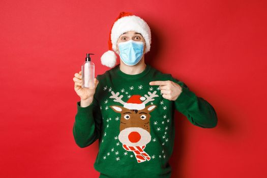 Concept of new year, coronavirus and social distancing. Portrait of cheerful man in santa hat, christmas sweater and medical mask, pointing finger at hand sanitizer, standing over red background.